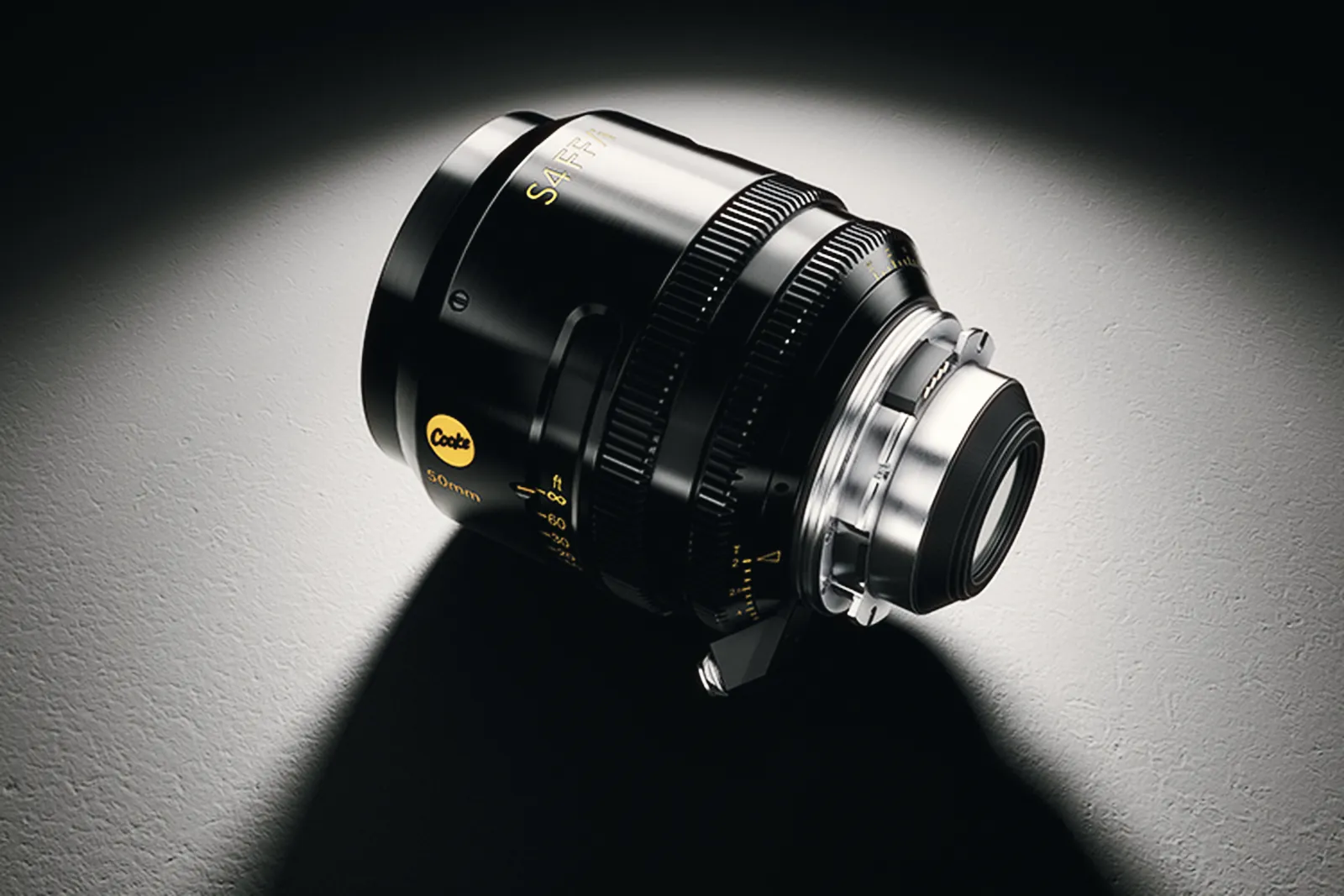 The Cooke S4 FF/i 50mm lying on its side under a spotlight, showing the gold Cooke badge, the focus scale in feet, two geared rings and the steel PL mount