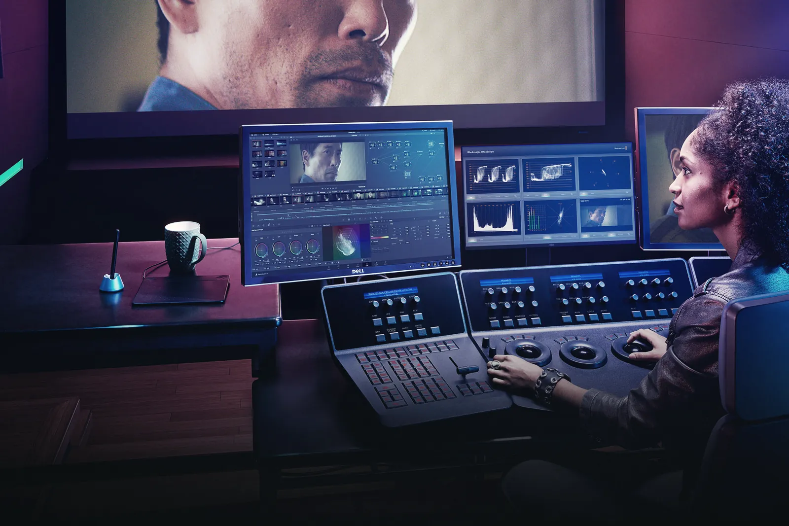 A colorist grading a close-up shot on a DaVinci Resolve control panel, with scopes and the Color page on two monitors and the image on a large reference screen