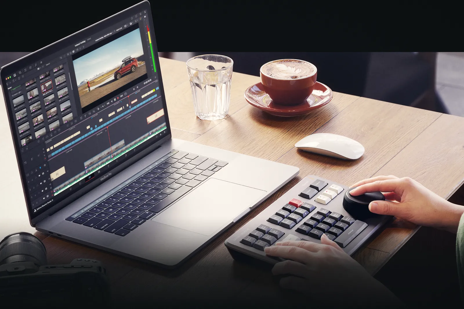 Hands on a DaVinci Resolve Speed Editor keyboard next to a laptop showing the Cut page, with a coffee cup and a glass of water on a wooden table