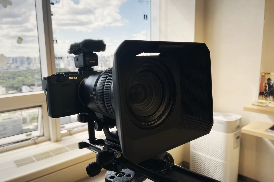 A black Nikon ZR cinema camera with a Nikon microphone and furry windscreen on top and a large zoom lens with a square matte box, on a slider by a window